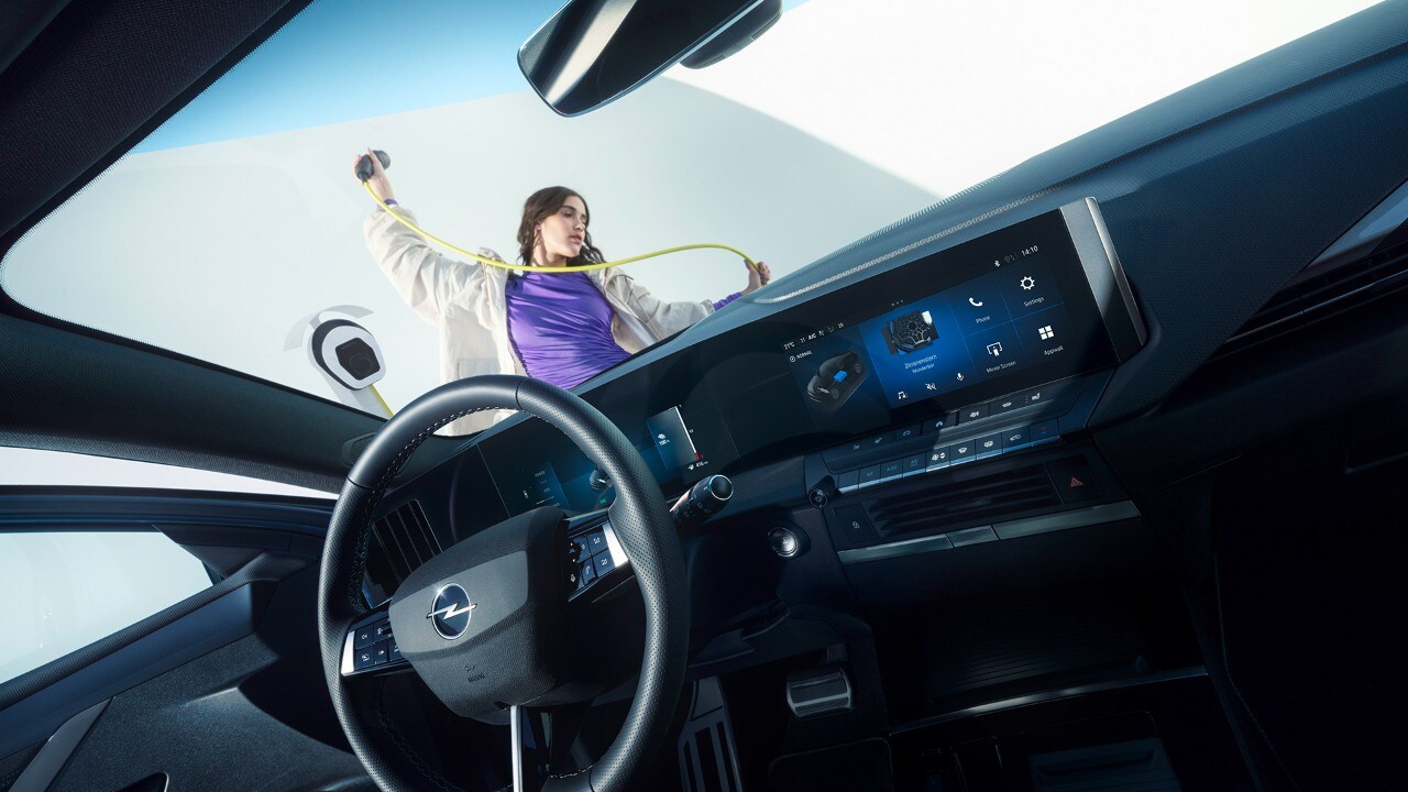 Vue intérieure d'une Opel Astra Electric depuis le siège passager avec une femme tenant un câble de recharge à l'extérieur.