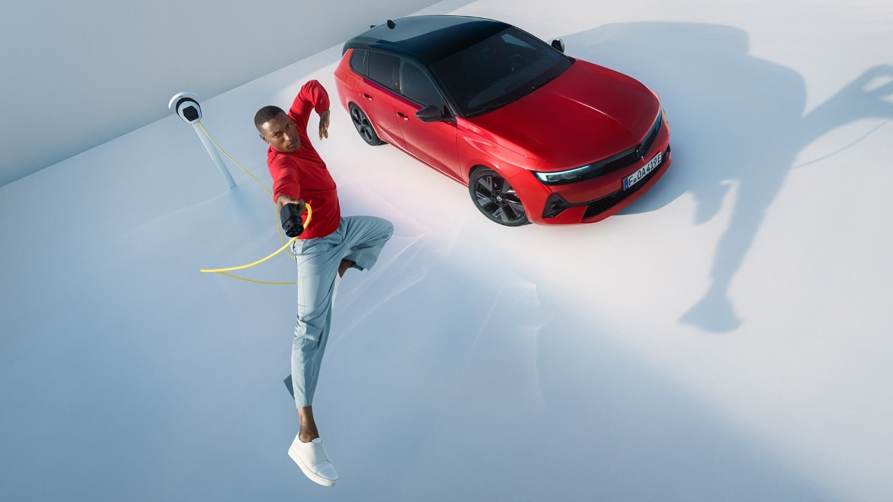 Homme sautant en l'air avec un câble de recharge pour véhicules électriques , avec une Opel Astra Sports Tourer Electric en arrière-plan.