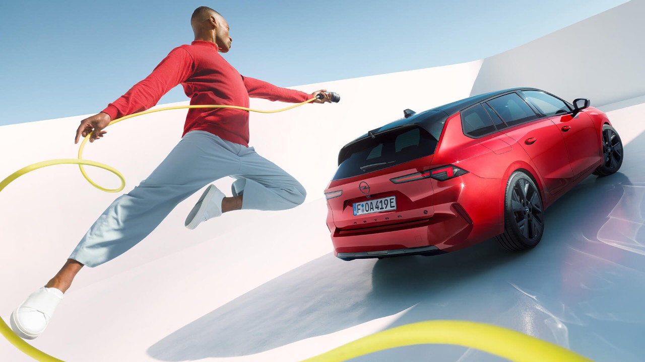 Homme sautant en l'air avec un câble de recharge pour véhicules électriques , avec une Opel Astra Sports Tourer Electric en arrière-plan.